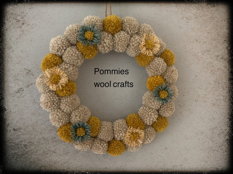 K&ouml;nnte beinhalten: Ein beige-gelber Pompon-Kranz mit kleinen Blumenakzenten. Der Kranz tr&auml;gt den Text "Pommies wool crafts" in der Mitte.
