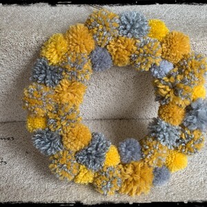 May include: A wreath made of yellow and gray pom-poms. The wreath is hanging on a wall.