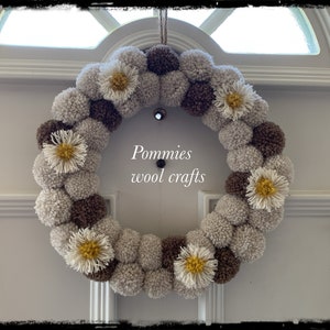 K&ouml;nnte beinhalten: Ein brauner und beiger Pompon-Kranz mit wei&szlig;en und gelben G&auml;nsebl&uuml;mchen. Der Kranz h&auml;ngt an einer wei&szlig;en T&uuml;r. Der Text "Pommies wool crafts" befindet sich in der Mitte des Kranzes.