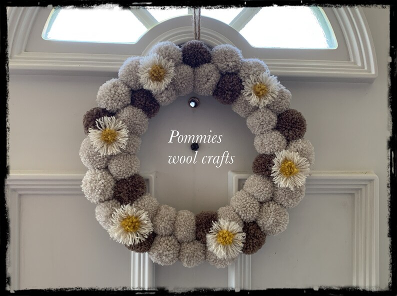 K&ouml;nnte beinhalten: Ein brauner und beiger Pompon-Kranz mit wei&szlig;en und gelben G&auml;nsebl&uuml;mchen. Der Kranz h&auml;ngt an einer wei&szlig;en T&uuml;r. Der Text "Pommies wool crafts" befindet sich in der Mitte des Kranzes.