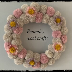 K&ouml;nnte beinhalten: Ein Kranz aus rosa und grauen Pompons mit gelben und violetten Blumenakzenten. Der Kranz tr&auml;gt den Text "Pommies wool crafts" in der Mitte.