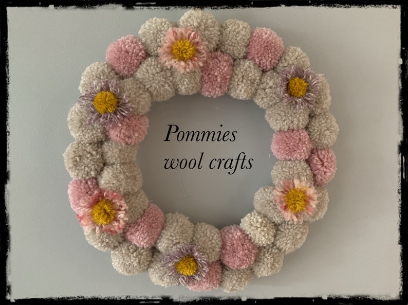 K&ouml;nnte beinhalten: Ein Kranz aus rosa und grauen Pompons mit gelben und violetten Blumenakzenten. Der Kranz tr&auml;gt den Text "Pommies wool crafts" in der Mitte.