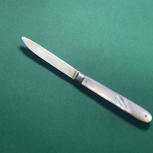 Puede incluir: Un cuchillo de bolsillo de plata con mango de nácar. La hoja está cerrada y el mango tiene un sutil patrón de remolino.