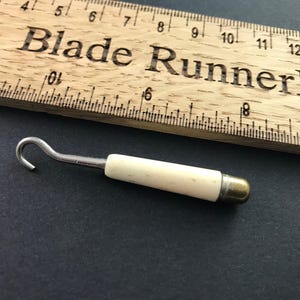 Può includere: Un utensile a gancio vintage con un manico color crema e un'estremità in ottone, appoggiato su una superficie scura. Un righello di legno con il testo "Blade Runner Fra" è sullo sfondo. Il gancio è in metallo.