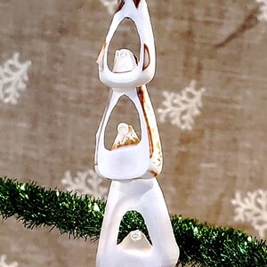 May include: A white seashell Christmas ornament with a star on top and a green ribbon. The ornament is made of three stacked seashells with holes in the centre. The ornament is hanging from a green garland.