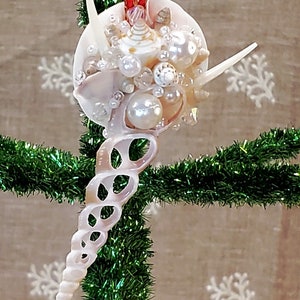 May include: A white seashell Christmas ornament with a spiral design and a cluster of smaller shells and pearls. The ornament is hanging on a green artificial Christmas tree.
