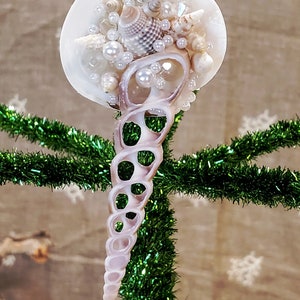 Puede incluir: Un adorno de concha blanca con una concha en espiral colgando de él. El adorno está decorado con perlas blancas y otras conchas.