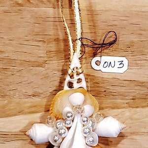 Puede incluir: Un adorno de ángel de conchas marinas con una cinta dorada y una etiqueta blanca que dice "ON 3". El ángel está hecho de varias conchas marinas y cuentas blancas.