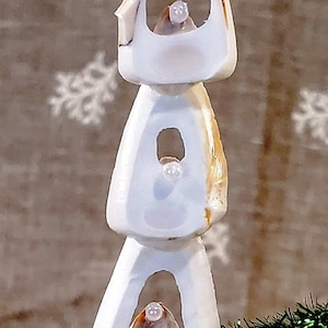 Può includere: Un ornamento per albero di Natale a forma di albero di Natale in conchiglia con una stella marina in cima e tre conchiglie bianche con dei buchi. L'ornamento è appeso a un albero di Natale artificiale verde.