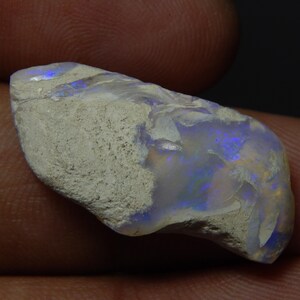 Op de afbeelding: Een ruwe, ongeslepen opaal edelsteen met een lichtgrijze, getextureerde buitenkant. De binnenkant vertoont iriserende flitsen van blauw en goud. De steen is onregelmatig van vorm en lijkt een natuurlijk, ongepolijst exemplaar te zijn.