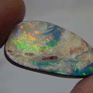 May include: A polished, irregular oval-shaped boulder opal gemstone featuring a dark base with vibrant flashes of green, blue, and orange play-of-color across its surface, set against a dark backing material with visible natural matrix inclusions.
