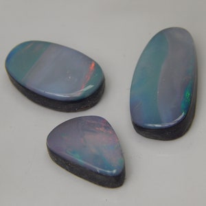 Peut inclure: Trois opales noires polies aux surfaces lisses et arrondies. Chaque pierre précieuse présente une couleur de base sombre avec des éclats iridescents de lumière bleue, violette et rouge. Les pierres sont façonnées en deux ovales et une forme triangulaire, toutes dotées d'un support sombre à la texture mate.