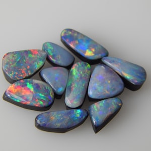 Peut inclure: Une collection de dix opales noires polies aux formes irrégulières. Chaque pierre précieuse présente une couleur de base sombre avec des éclats vibrants de lumière irisée rouge, verte, bleue et jaune réfléchis sur les surfaces lisses.