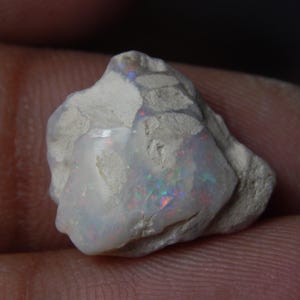 Puede incluir: Una piedra preciosa de ópalo en bruto, sin cortar, con una base blanca y destellos iridiscentes de rojo, verde y azul. El ópalo se sostiene en una mano, mostrando su textura natural y sus colores vibrantes.
