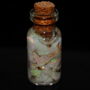 Könnte beinhalten: Eine kleine, klare Glasflasche gefüllt mit schillernden Opalfragmenten. Die Flasche ist mit einem Korken verschlossen. Die Opale zeigen grüne, orange und gelbe Blitze auf weißem Hintergrund. Die Flasche steht vor einem schwarzen Hintergrund.