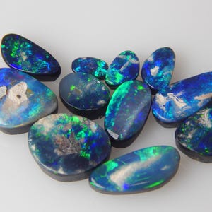 May include: A collection of ten polished black opals featuring vibrant flashes of blue and green color play. Each gemstone has a smooth, rounded surface with irregular shapes and varying sizes, displaying natural inclusions and dark base tones.