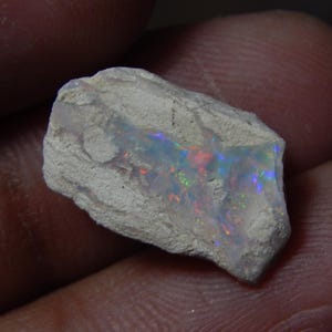 Op de afbeelding: Een ruwe, ongeslepen opaal edelsteen met een lichtgrijze buitenkant. De opaal vertoont een levendig kleurenspel, met flitsen van rood, blauw en groen zichtbaar in de steen. De edelsteen wordt in een hand gehouden.