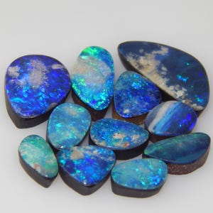 May include: A collection of ten polished loose opals featuring vibrant blue and green flashes of color. Each gemstone has a dark, opaque backing and an irregular, rounded shape, displaying a play-of-color across their smooth, reflective surfaces.
