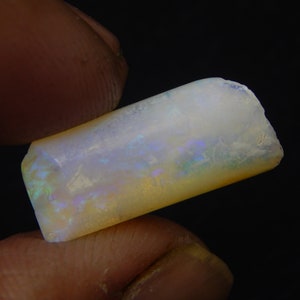 May include: A rough-cut opal gemstone, rectangular in shape, displaying a milky white base with iridescent flashes of blue, green, and yellow. The opal is held against a dark backdrop, highlighting its natural, unpolished texture and colour variations.