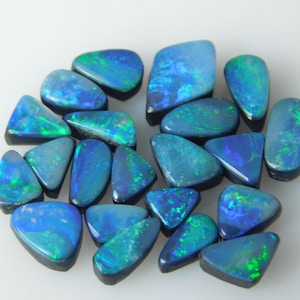 May include: A collection of loose, polished black opals in various geometric shapes. Each gemstone features a dark base color with vibrant flashes of blue and green play-of-color, displaying a smooth, reflective surface and distinct edges.
