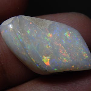 Peut inclure: Une pierre d'opale polie et irisée. La pierre a une couleur de base blanc laiteux avec des éclairs de vert, d'orange et de bleu. L'opale est de forme irrégulière avec une surface lisse et brillante.
