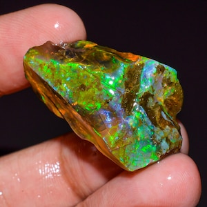 May include: A raw, unpolished opal gemstone displaying vibrant flashes of neon green, electric blue, and warm orange play-of-color across its irregular, translucent surface. The stone features deep, earthy brown inclusions and a glossy, wet texture.