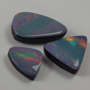 May include: Three polished black opal gemstones with iridescent color flashes of red, green, and yellow. The opals have a dark base color with a glossy finish. The shapes are teardrop and triangular.