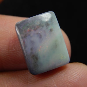 May include: A rectangular opal gemstone with rounded corners. The stone displays a mix of colors, including blue, green, and hints of red and brown. The opal has a smooth, polished surface and a semi-translucent appearance.