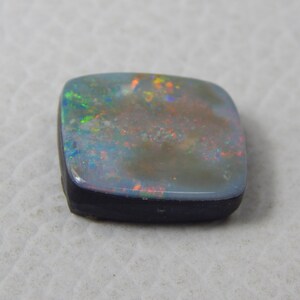 May include: A square-cut opal gemstone with a dark base and a shimmering, iridescent surface. The opal displays a play of color with flashes of green, orange, and blue. The gemstone is set against a neutral background.