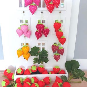 Op de afbeelding: Een verzameling stoffen aardbei-ornamenten in rood, roze en geel met witte stippen en groene viltblaadjes. Sommige hangen aan een wit houten huisvormig displayrek, terwijl andere in een bakje aan de onderkant verspreid liggen.