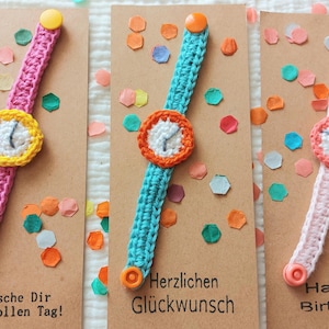 Könnte beinhalten: Drei gehäkelte Armbänder auf braunem Karton, jedes mit einem runden Zifferblatt mit Zeigern und einem Druckknopfverschluss. Die Bänder sind rosa, türkis und hellrosa, mit gelben, orangen und rosa Druckknöpfen und Konfetti.
