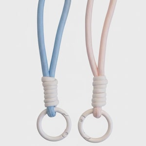 May include: Two keychains, one baby blue and one light pink, each with a white ring and a matching cord. The cords are attached to the rings with a white, tightly wound knot. The background is a solid, neutral color.