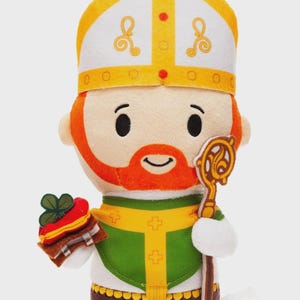 May include: A plush toy of a saintly figure, featuring a white and gold mitre, orange beard, and green vestments. The figure holds a staff and a stack of what appears to be a cake with a shamrock. The toy has a smiling expression.