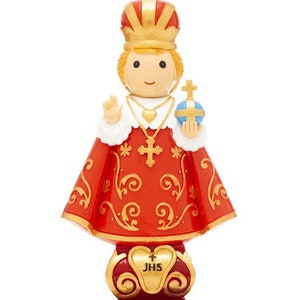May include: A small figurine of a child king wearing a red and gold robe with a crown and holding a globe. The figurine is standing on a base with the inscription "JHS".