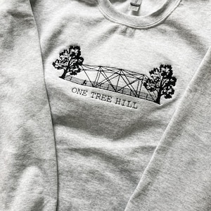 Puede incluir: Sudadera gris con un dise&ntilde;o bordado en negro de un puente con dos &aacute;rboles a cada lado y el texto "ONE TREE HILL".