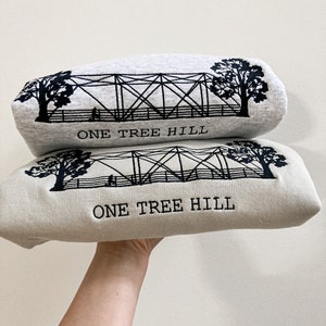 Puede incluir: Dos sudaderas grises con bordado negro de un puente y &aacute;rboles. El texto "ONE TREE HILL" est&aacute; bordado en ambas sudaderas.