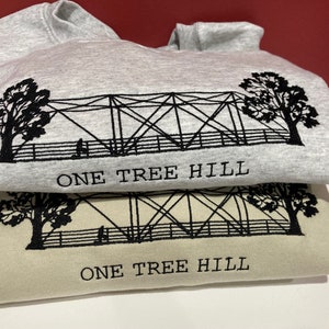 Puede incluir: Dos sudaderas con un dise&ntilde;o bordado en negro de un puente y &aacute;rboles con el texto "One Tree Hill" en cada una. La sudadera de arriba es gris y la de abajo es beige.