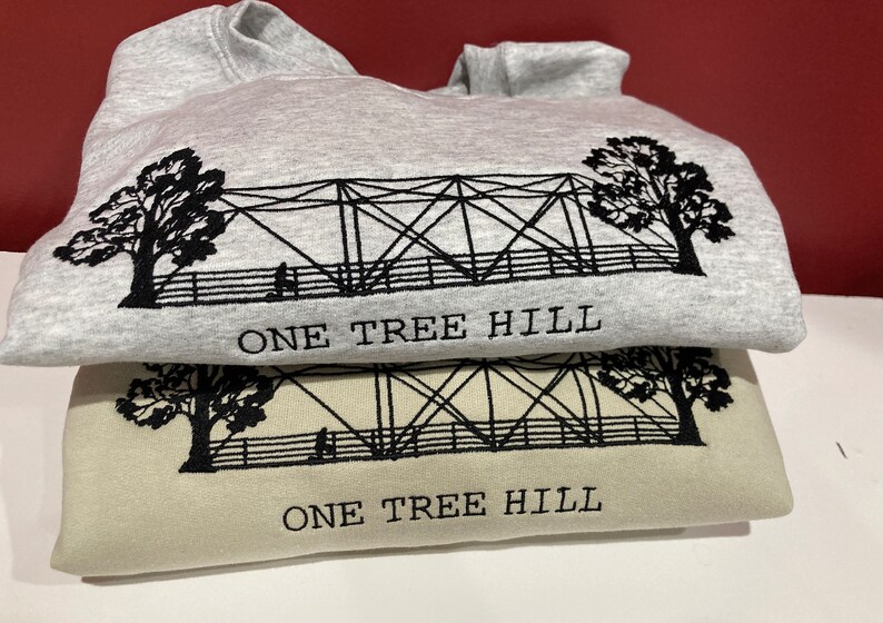 Puede incluir: Dos sudaderas con un dise&ntilde;o bordado en negro de un puente y &aacute;rboles con el texto "One Tree Hill" en cada una. La sudadera de arriba es gris y la de abajo es beige.