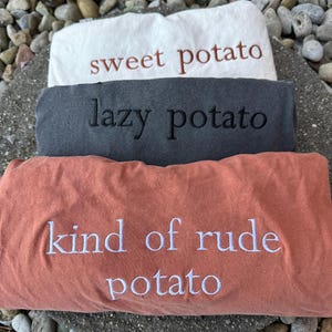 May include: Three folded sweatshirts in white, charcoal gray, and coral. The white sweatshirt has "sweet potato" embroidered in brown. The gray sweatshirt has "lazy potato" embroidered in black. The coral sweatshirt has "kind of rude potato" embroidered in white.