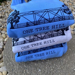Puede incluir: Tres sudaderas azules y grises apiladas una encima de la otra. Cada sudadera tiene un gr&aacute;fico negro de un puente con el texto "One Tree Hill" impreso debajo.