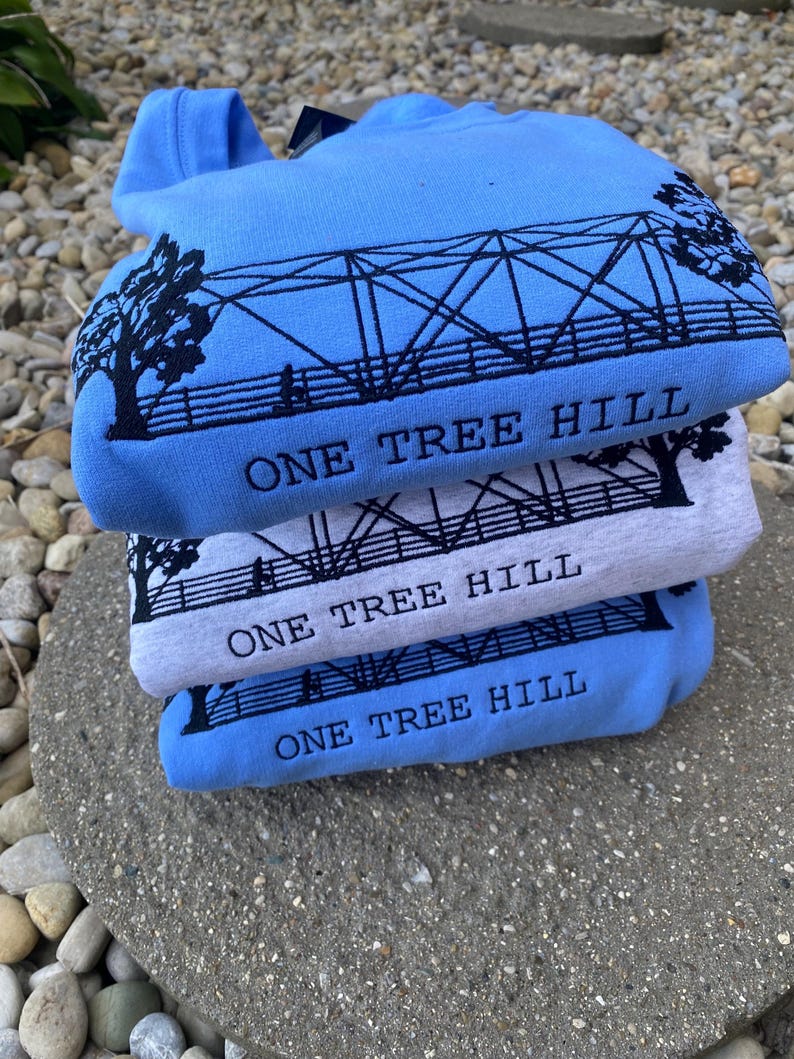 Puede incluir: Tres sudaderas azules y grises apiladas una encima de la otra. Cada sudadera tiene un gr&aacute;fico negro de un puente con el texto "One Tree Hill" impreso debajo.