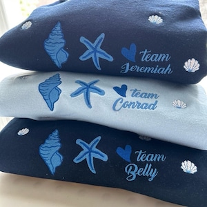 May include: Three folded sweatshirts in navy, light blue, and black. Each features embroidered seashell, starfish, and heart designs, along with the text "Team Jeremiah", "Team Conrad", and "Team Belly".