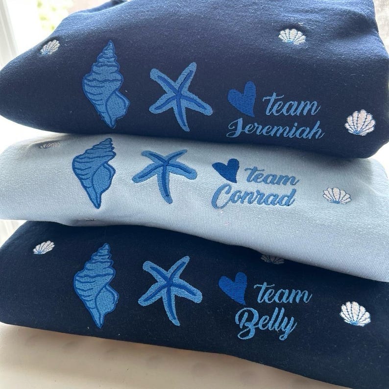May include: Three folded sweatshirts in navy, light blue, and black. Each features embroidered seashell, starfish, and heart designs, along with the text "Team Jeremiah", "Team Conrad", and "Team Belly".