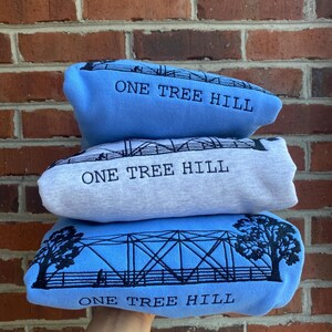 Puede incluir: Tres sudaderas azules apiladas una encima de la otra. Cada sudadera tiene un dise&ntilde;o bordado en negro de un puente con el texto "One Tree Hill" debajo.