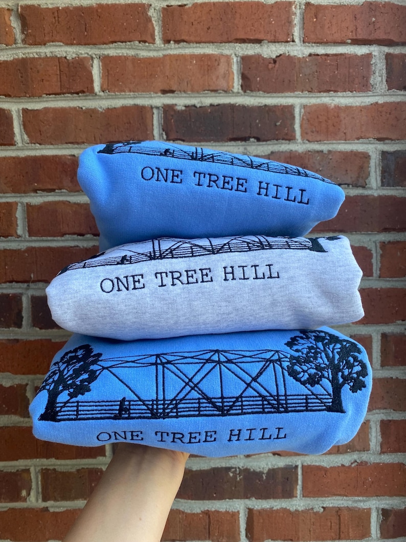 Puede incluir: Tres sudaderas azules apiladas una encima de la otra. Cada sudadera tiene un dise&ntilde;o bordado en negro de un puente con el texto "One Tree Hill" debajo.