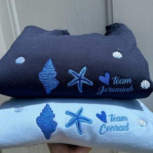 May include: Two sweatshirts, one navy blue and one light blue, are stacked. Both have embroidered blue seashell, starfish, and heart designs. The navy sweatshirt says "Team Jeremiah," and the light blue one says "Team Conrad."