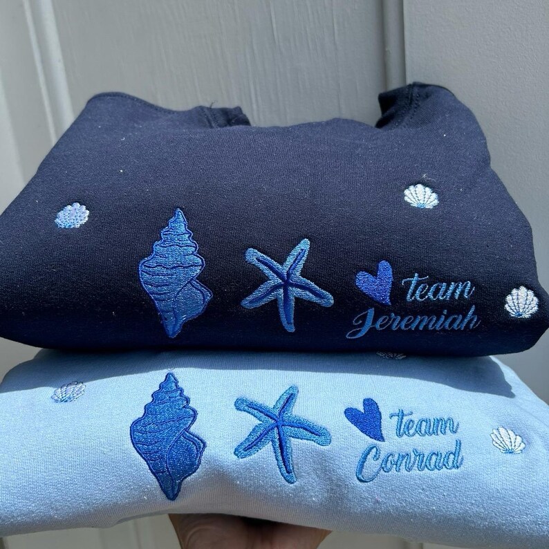 May include: Two sweatshirts, one navy blue and one light blue, are stacked. Both have embroidered blue seashell, starfish, and heart designs. The navy sweatshirt says "Team Jeremiah," and the light blue one says "Team Conrad."