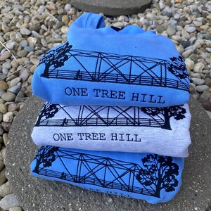 Puede incluir: Tres camisetas dobladas en tonos azules y grises. Cada camiseta presenta una silueta negra de un puente con el texto "One Tree Hill" impreso debajo.