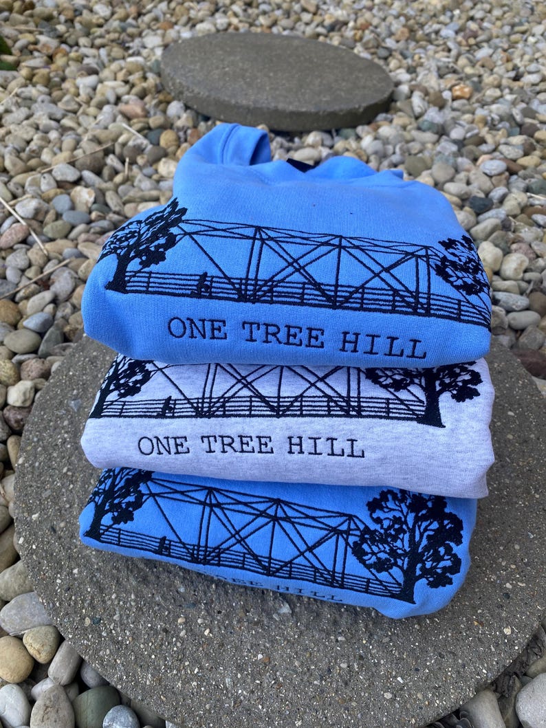 Puede incluir: Tres camisetas dobladas en tonos azules y grises. Cada camiseta presenta una silueta negra de un puente con el texto "One Tree Hill" impreso debajo.