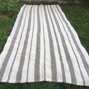 May include: A long, white and brown striped rug with a woven texture. The rug is laid out on a grassy surface.